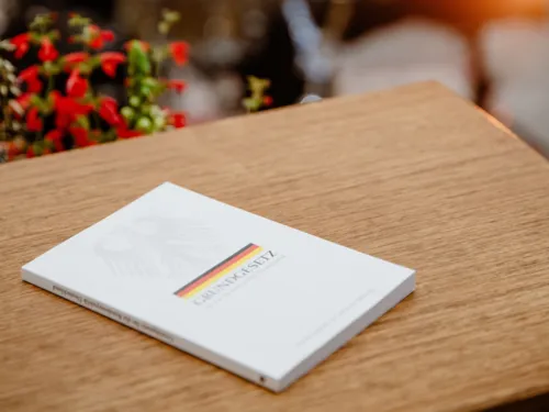 Kleine Taschenbuchausgabe des Grundgesetzes auf einem hellen Holztisch. Im Bildhintergrund außerhalb des Fokus ein Blumenstrauß.