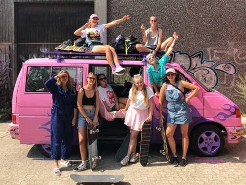 ein pinkfarbener Kleinbus. Davor, darin und darauf acht junge Frauen mit Skateboards