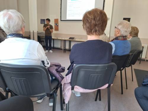 Eine Gruppe Senior*innen sitzt im Halbkreis und hört einem Referenten beim Vortrag zu.