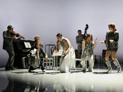 Auf einer Bühne im Hintergrund ein Violinist, ein Klavierspieler, ein Saxofonist und ein Bassist, davor singen tanzend drei Frauen und ein Mann.