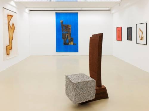 Zu sehen ist ein Galerie-Innenraum. Ausgestellt sind Arbeiten des Künstlers Hartmut Stielow. An den drei sichtbaren Wänden hängen groß- und mittelformatige Bilder. In der Raummitte ist eine geometrische Plastik aus rostigem Stahl und einem Grafit-Block zu sehen.