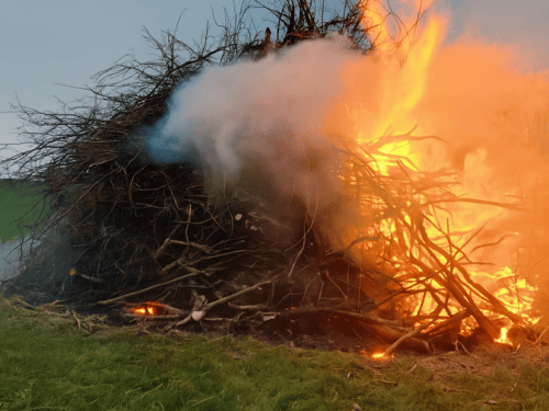 Zu einem großen Haufen aufgeschichtete dicke und dünnere Äste auf einem Feld, die brennen.