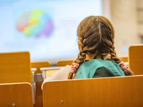 Sitzendes Kind in einem Hörsaal mit geflochtenen Zöpfen von hinten aufgenommen.