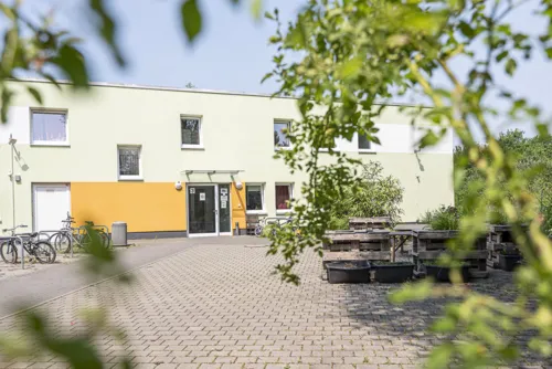 Ein flaches, zweistöckiges Gebäude mit einigen Fenstern und einem überdachten Eingang. Das Gebäude ist mintgrün und hell-orange gestrichen. Vor dem Gebäude einige Hochbeete, Bänke und Tische.
