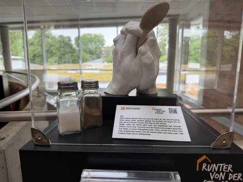 Zu sehen ist ein Foto einer Installation: Zwei Gipshände auf einem Sockel stehend halten einen Holzlöffel, daneben stehen ein Salz- und Pfefferstreuer.