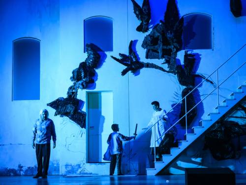 Auf einer Bühne ist vor einer Häuserfassade eine Treppe zu sehen. Links davon ist ein Mann Zeuge, wie ein Kind einer Frau, die auf der Treppe steht, einen Dolch überreicht.