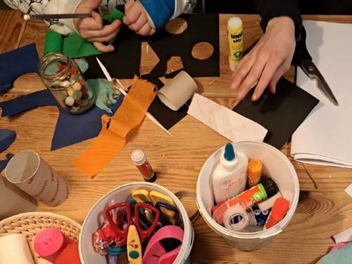 Auf einem Tisch liegen verschiedene Bastelmaterialien. Hände von einem Kind und einer erwachsenen Person arbeiten damit.