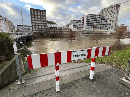 Ein rot-weiß-gestreiftes Schild, dahinter Wasser und ein Gebäudekomplex. Links eine Brücke,