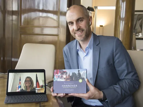 Ein Mann hält einen Reiseführer in den Händen und lächelt in die Kamera, neben ihm steht ein Laptop, auf dem eine Frau in einer Videokonferenz zu sehen ist, auch sie lächelt