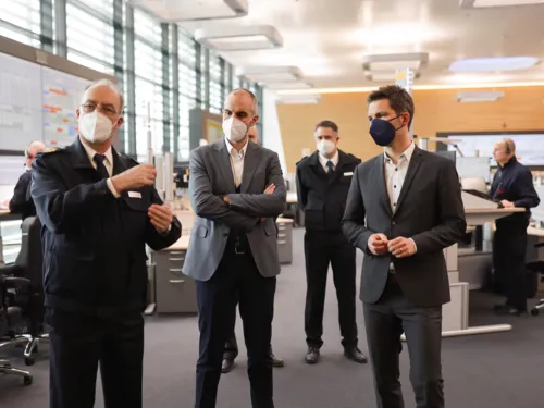 Drei Männer stehen in einem Raum und unterhalten sich, im Hintergrund stehen weitere Personen an Schreibtischen