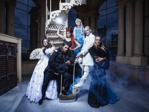 Zu sehen sind Musicaldarsteller und Musicaldarstellerinnen in verschiedenen Kostümen, die vor einer weißen Wendeltreppe stehen.