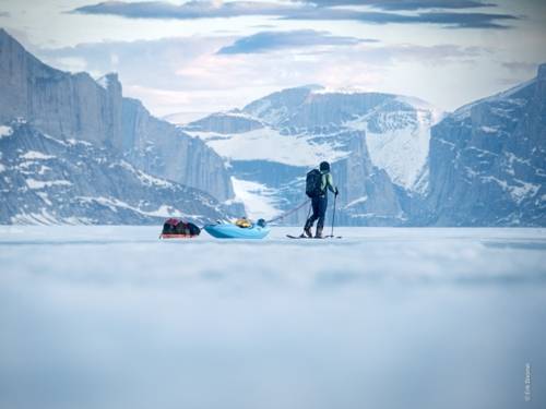 Zu sehen ist ein Film Still: Eine Eislandschaft wird im Hintergrund von Bergen umsäumt, im Vordergrund ist eine Person in passender Ausrüstung zu sehen, die sich über einen Eisboden bewegt.