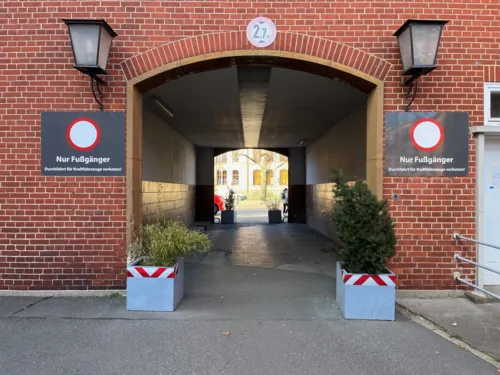 Ein Torbogen mit einem Durchgang in einem roten Klinkergebäude. Rechts und links des Bogens stehen Pflanztröge, darüber sind am Haus Schilder "Nur Fußgänger" angebracht. Hinter dem Durchgang ist Fußweg und der Ausschnitt eines weiteren Gebäudes zu sehen.