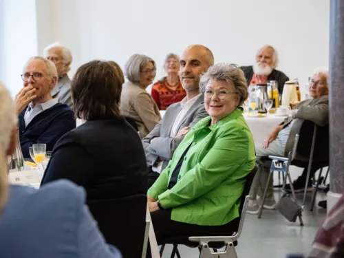 Menschen sitzen an Tischen, in der Mitte eine Frau und ein Mann, die in die Kamera sehen.
