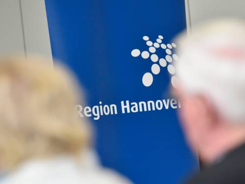 Auf einem Banner ist das Logo der Region Hannover in Weiß auf blauem Grund zu sehen. Davor sitzen Menschen.