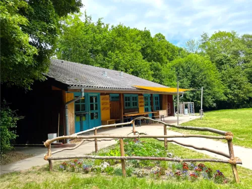 Eine Holzhütte mit blauen Fenster- und Türelementen. Dahinter liegen Bäume und davor eine Wiese mit einem umzäunten Blühbeet.