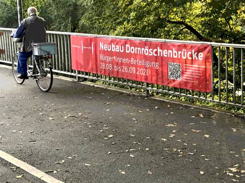 Banner auf einer Brücke, auf dem "Neubau Dornröschenbrücke" und ein QR-Code stehen. Eine Person auf einem Fahrrad fährt vorbei.
