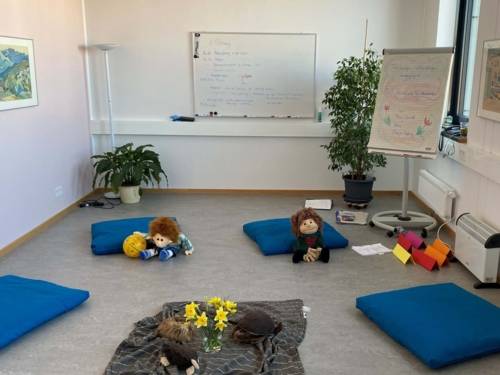 Ein Beratungsraum. Auf dem Boden liegen Decken, Kissen, Blumen und Puppen. Im Hintergrund eine Tafel und ein Whiteboard.