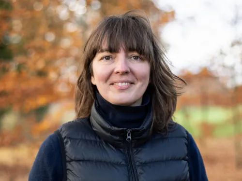 Eine dunkelhaarige Frau in schwarzem Pulli und Weste steht vor einem herbstlichen Baum.