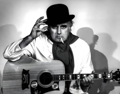 Mann mit Hut, Halstuch, Zigarette im Mund und Gitarre auf dem Schoß