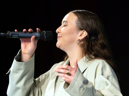 Eine Frau in einem hellen Mantel singt mit geschlossenen Augen in ein Mikrofon. Die Frau ist im Profil zu sehen, der Bildhintergrund ist schwarz.