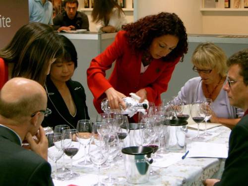 Zwei Männer und zwei Frauen sitzen an einem Tisch, zwei Frauen in Messeuniformen gießen ihnen Wein in bereitstehenden Gläsern ein.