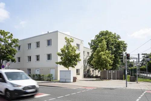 Ein beiges, dreistöckiges Gebäude mit Flachdach. Davor einige Bäume und eine befahrene Straße.