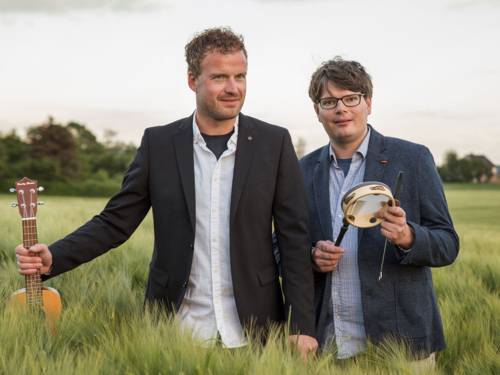 Zwei Männer mit Gitarre bzw. kleiner Trommel auf einer Wiese mit hüfthohem Gras