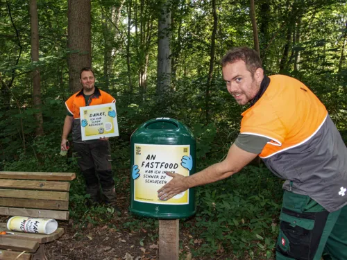 Zwei M&auml;nner, die einen Aufkleber an einem M&uuml;lleimer im Wald anbringen.