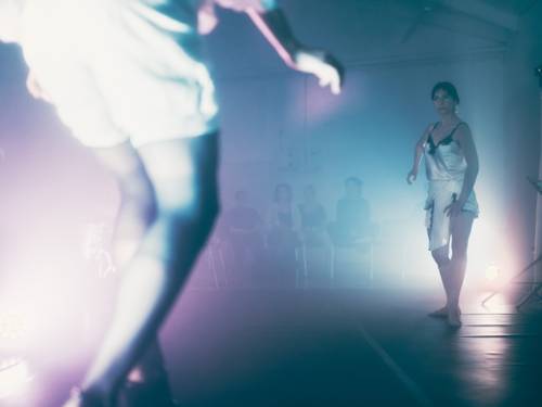 Zu sehen sind zwei Perfomerinnen auf einer ebenerdigen Bühne. Im Hintergrund sich Zuschauende zu erkennen. Das ganze Bild ist in einem weichen Licht aus Pink und Blau gehalten.