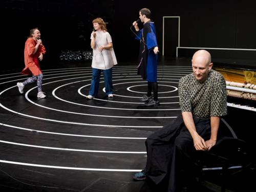 Zwei Schauspielerinnen und ein Schauspieler singen auf einer Bühne in Mikrofone, davor sitzt ein Mann an einem Klavier.