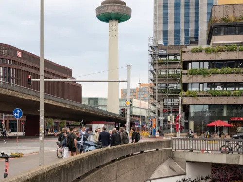 Menschen stehen auf einem Platz, links eine Kreuzung unter einer Hochstra&szlig;e, hinten in der Mitte ein gro&szlig;er Funkturm, rechts hinten ein Geb&auml;ude, vorn rechts eine tiefere Untergeschossebene.
