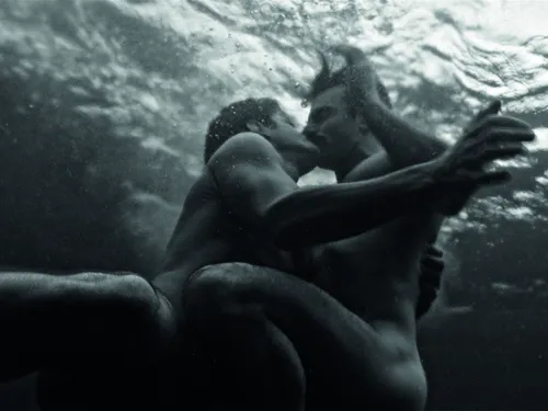 Das Schwarz-Weiß-Bild zeigt zwei Personen unter Wasser in einer innigen Umarmung. Die Aufnahme wurde aus einer Perspektive unterhalb der Wasseroberfläche fotografiert, wodurch Lichtreflexe und die Bewegung des Wassers eine traumartige Atmosphäre erzeugen. Die Körper der beiden Personen sind nur teilweise sichtbar und verschmelzen mit den weichen Kontrasten der Umgebung zu einer poetischen, künstlerischen Komposition.