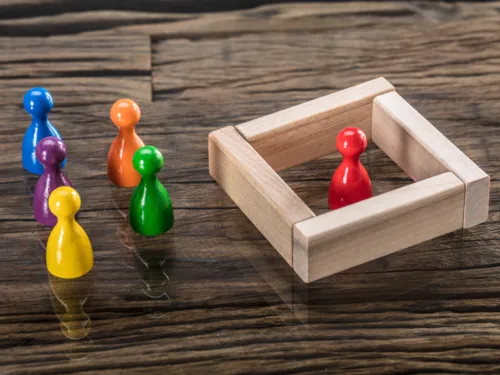 Links stehen bunte Spielfiguren auf einem Tisch. Rechts daneben steht eine Figur in einer anderen Farbe, die durch Holzplatten eingesperrt ist.