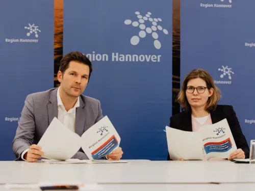 Ein Mann un deine Frau sitzen an Tischen und halten jeweils ein Konzeptpapier in den Händen.