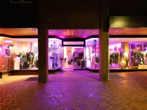 Eine Schaufensterzeile bei Nacht in lila Neonlicht. Im Ladengeschäft sind hauptsächlich Kleidungsstücke zu sehen.