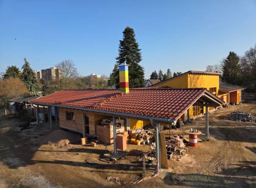 Ein Gebäude mit einem Geschoss, roten Ziegeln und einem farbig gestreiften Schornstein von seitlich oben fotografiert. Drum herum ein sandiger Weg, Steinhaufen und Baumaterialien wie Bretter udn Eimer.