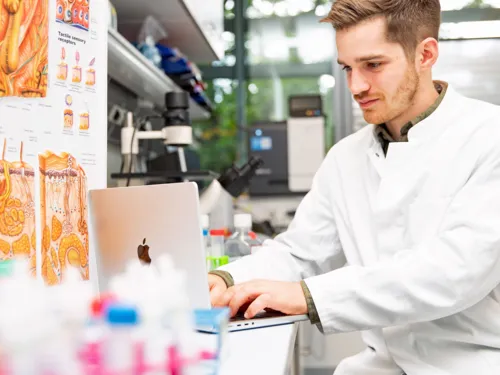 Ein Mann im weißen Kittel sitzt im Labor am Computer neben einem Plakat, das den Aufbau der Haut darstellt.