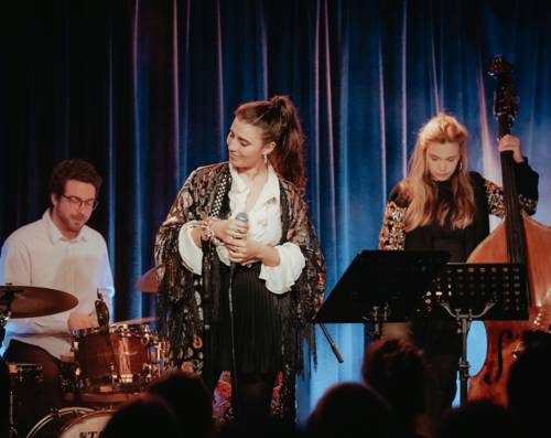 Ein Mann an einem Schlagzeug und zwei Frau mit Mikro bzw. Kontrabass auf einer Bühne