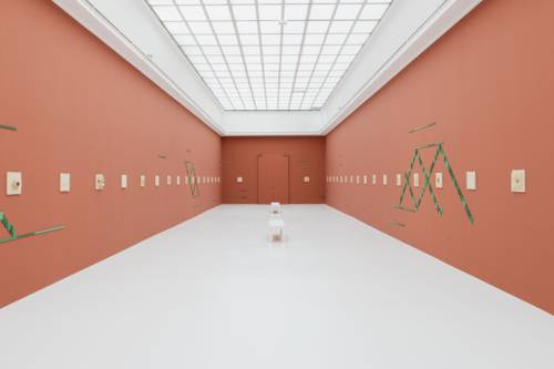 Ein Stuhl in einem Ausstellungsraum, an dessen Wände Kunst hängt und klebt