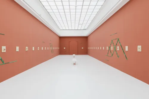 Ein Stuhl in einem Ausstellungsraum, an dessen Wände Kunst hängt und klebt