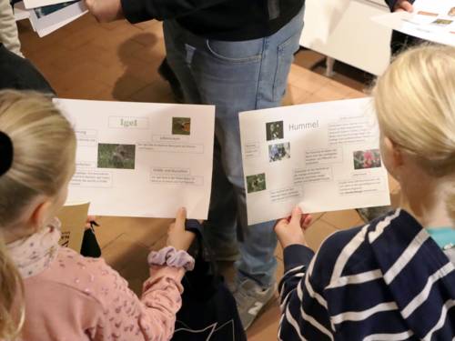 Zwei Mädchen halten jeweils ein Blatt Papier mit Informationen: Links über den Igel, rechts über die Hummel.