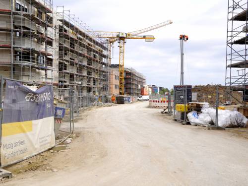 Links ein Rohbau mit einem Baugerüst, vorn ein Bauzaun, weiter hinten ein Kran. Am Bau führt eine mit sand überdeckte Baustraße vorbei.