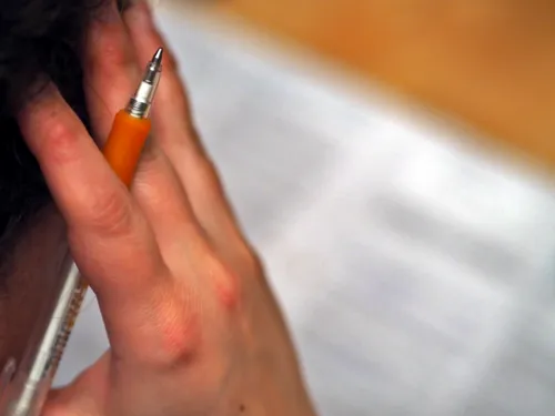 Eine Frau hat die rechte Hand nachdenklich an den Kopf gelegt, zwischen den Fingern klemmt ein Kugelschreiber. Vor ihr liegt ein Schriftstück.