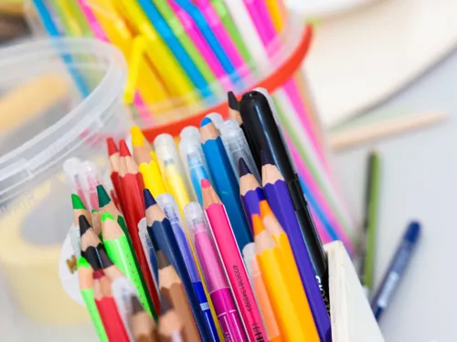 Verschiedene Stift stehen in einem Plastikbecher, dahinter stehen bunte Strohhalme in einem weiteren Plastikbecher.