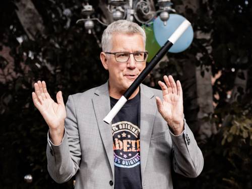 Mann mit kurzem Haar und Brille in T-Shirt und Jackett, zwischen seinen Händen schwebt ein Zauberstab.