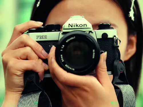 Das Bild zeigt eine Person, die eine analoge Spiegelreflexkamera vom Typ Nikon FM2 vor das Gesicht hält und durch den Sucher fotografiert. Die Kamera steht im Mittelpunkt der Aufnahme und wird von beiden Händen umfasst. Durch die Nahaufnahme sind Details des Kameragehäuses und des Objektivs deutlich sichtbar, während der Hintergrund unscharf bleibt. Die warmen Farben und die geringe Tiefenschärfe verleihen dem Bild eine konzentrierte, fotografische Atmosphäre.