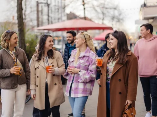 im Vordergrund sind vier Frauen in einer Reihe, die ein Getränk in der Hand halten und sich unterhalten. Im Hintergrund sind weitere personen zu sehen.