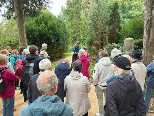 Menschen stehen umringt von Bäumen und Büschen im Halbkreis einem Mann zugewandt, der von einem Papier abliest.