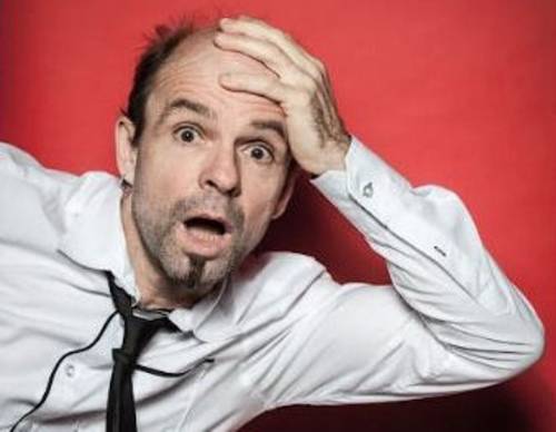 Mann mit kurzem Resthaar, Dreitagebart, hochgezogenen Augenbrauen und geöffnetem Mund hält sich die linke Hand an den Kopf.